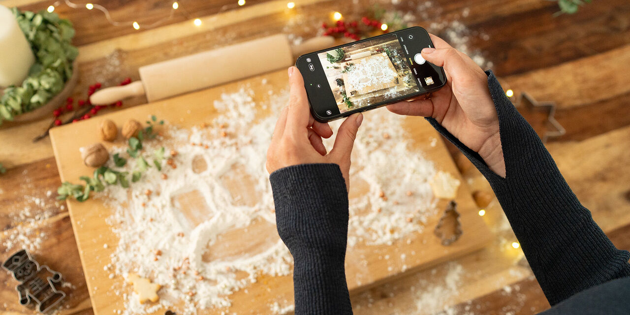 Je vidět velký dřevěný vál posypaný moukou a vánočně vyzdobený. V popředí jsou vidět dvě ruce, které drží chytrý telefon ve vodorovné poloze a fotografují s ním vál..