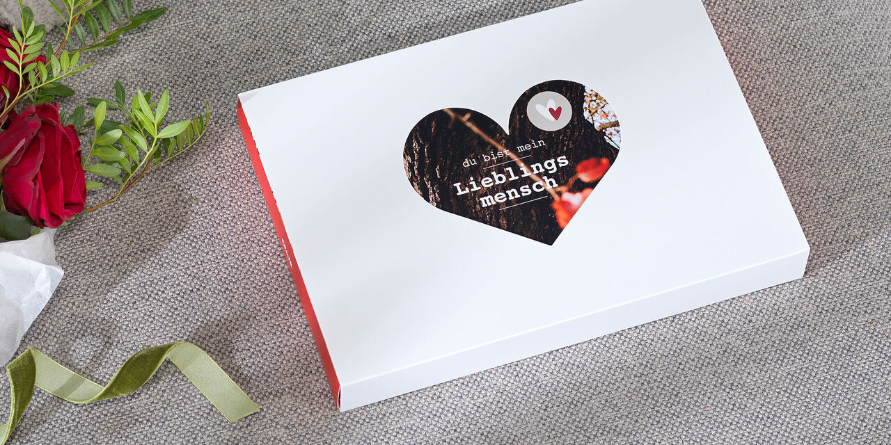 Zabalená valentýnská dárková krabička s fotkou leží na pohovce. Část víka krabice je vidět ve výřezu srdce. Říká „Oblíbená osoba“.
