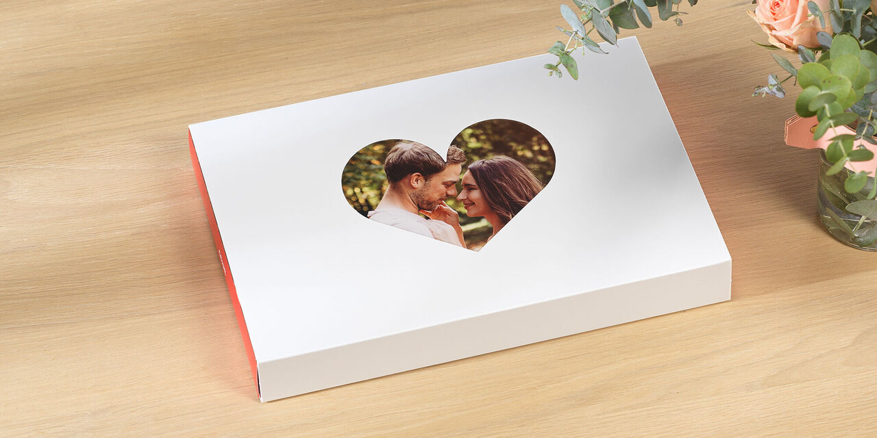 Uzavřená valentýnská krabička s fotkou leží na stole. Fotografie páru je vidět ve výřezu srdce. Vedle krabice jsou květiny a croissanty.