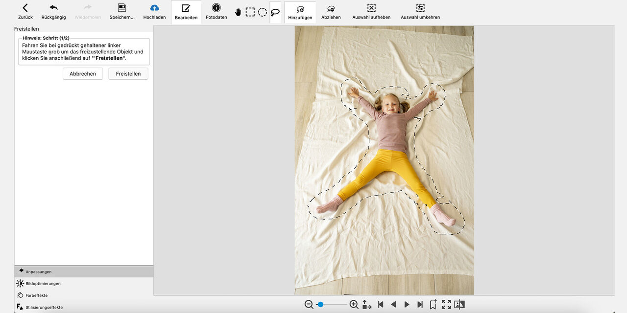 Snímek obrazovky z programu CEWE fotosvět: Dívka leží na podlaze na světlé rozprostřené dece a usmívá se do kamery. Dívka má natažené obě ruce a nohy daleko od sebe. Obrys těla je zhruba načrtnut přerušovanou čarou.